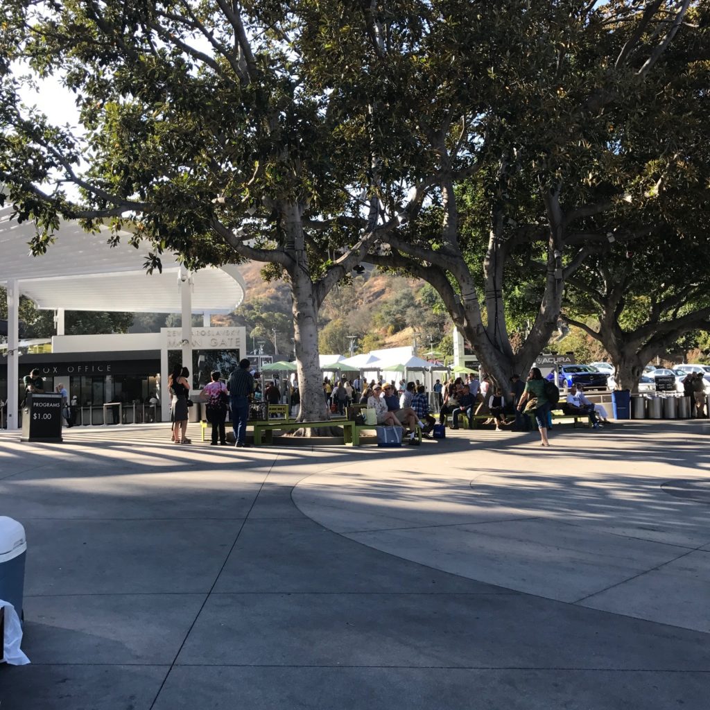 Image of the Hollywood Bowl entrance grounds with people standing under shade trees and the box office in the background.