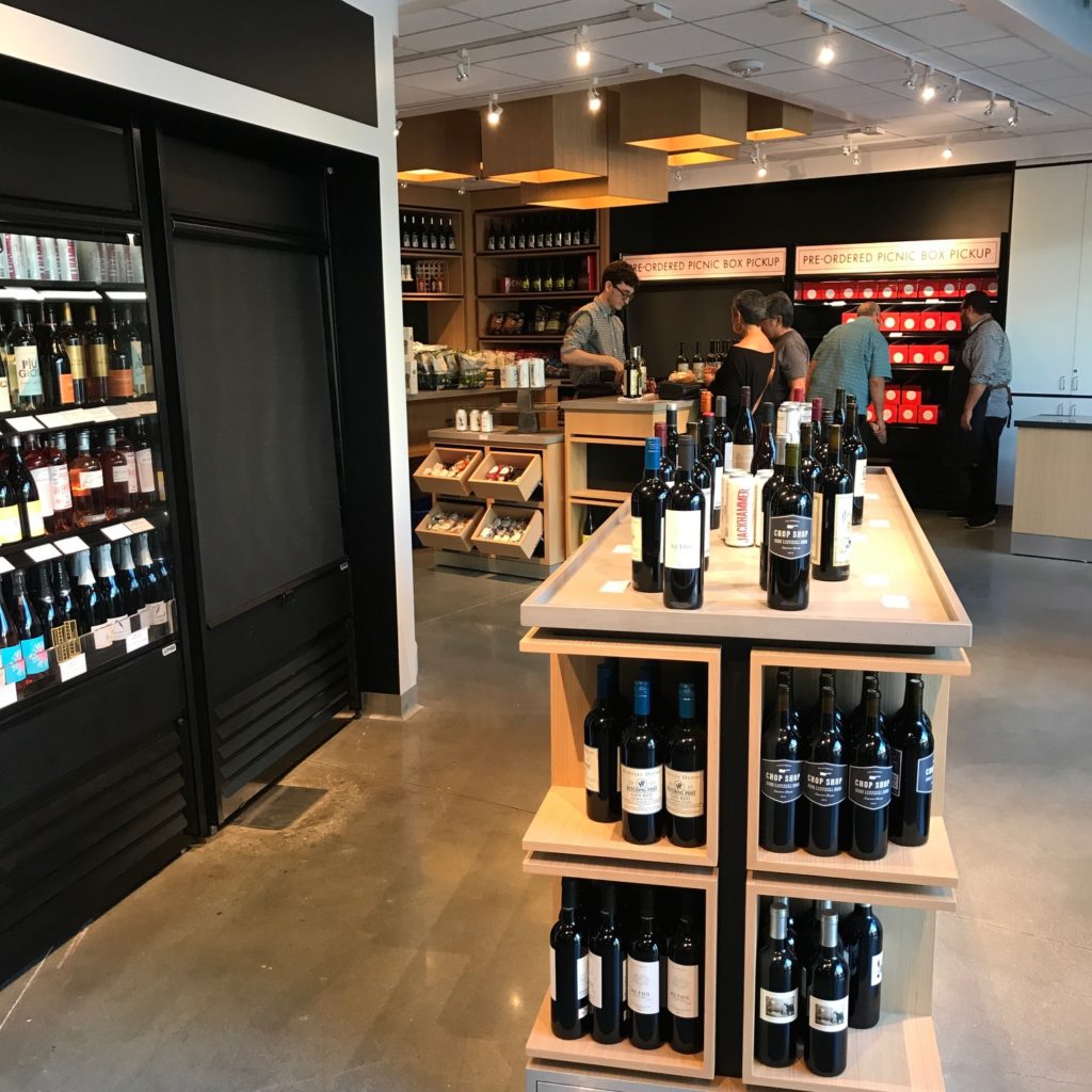 Image of the Hollywood Bowl Marketplace with open-shelf wine cooler to the left and display of wine bottles in the foreground. In the background an employee is checking out some shop patrons while another employee assists a man in the pre-order pickup area.